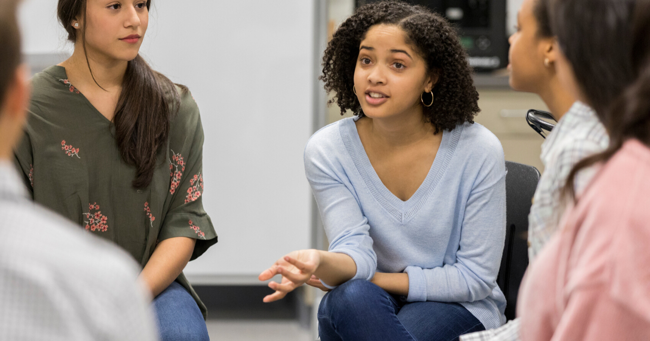 A group of teen girls sit in a circle and talk. 