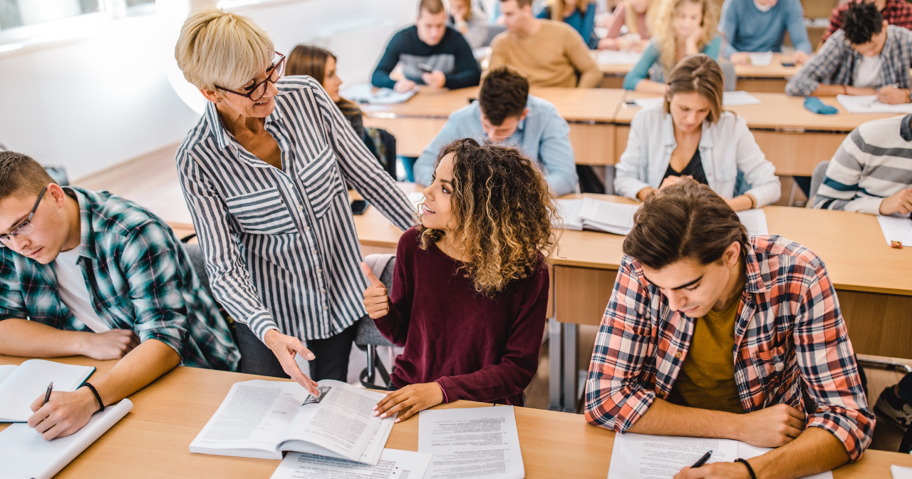 A teacher talks encouragingly to a student in a full classroom. 