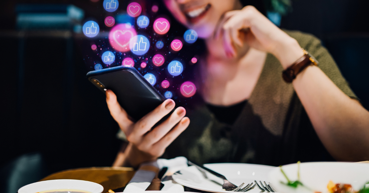A woman looks at her phone while eating a meal and images of social media icons float out from the screen. 