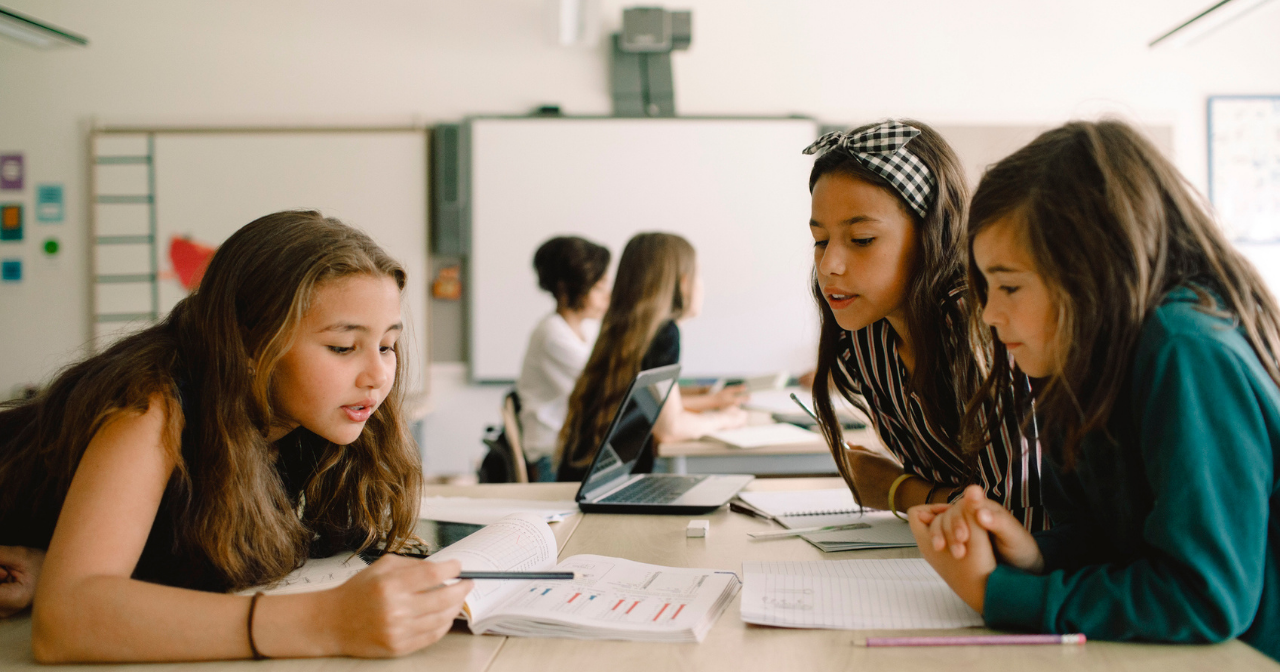 Three young girls sit at a desk in a classroom and discuss a piece of paper in front of them. 