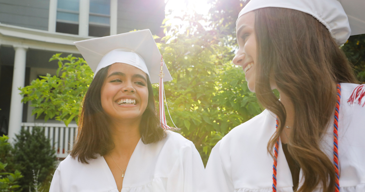 Two girls wearing white high school graduation gowns and smile. 