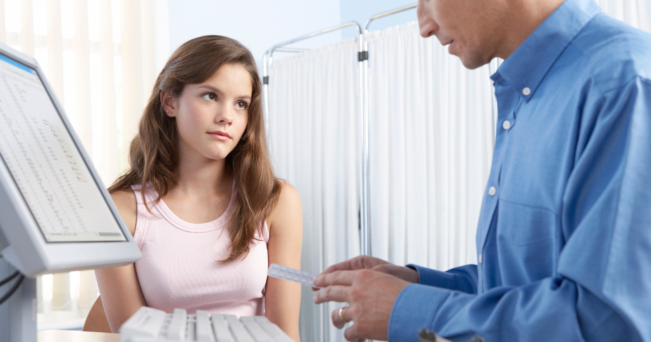A male provider talks to a young teen about birth control while holding a packet of birth control pills. 