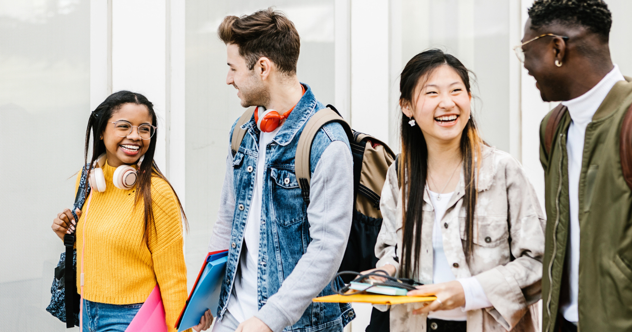 A group of four smiling teens walk together with their bookbags. 