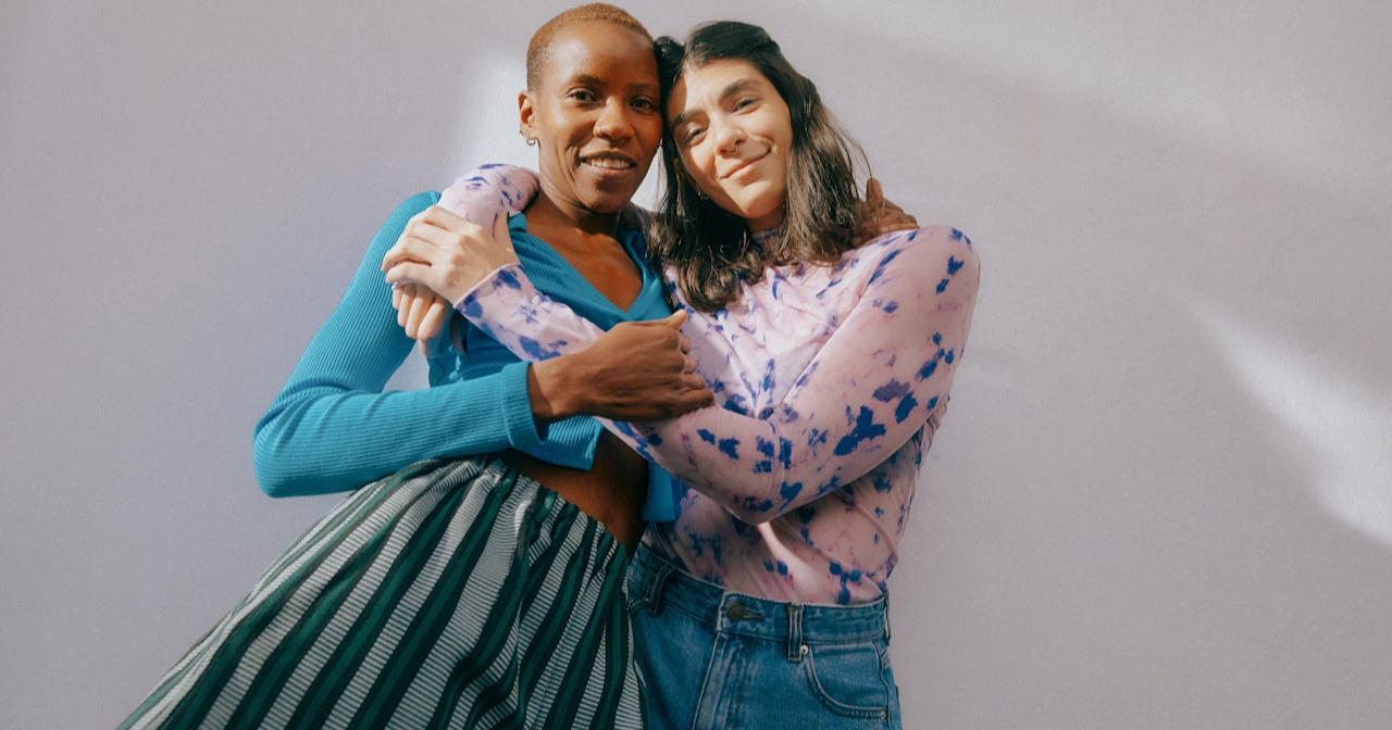 Two women stand against a blank wall and side hug each other. 