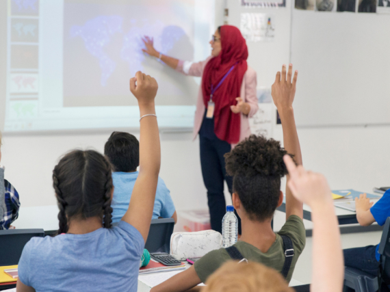 A stands teacher at the front of a classroom while students raise their hands. 