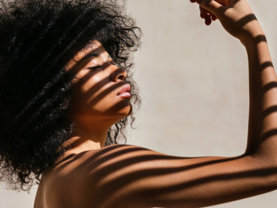 A Black woman curls her bicep while the sun shines through blinds making shadows across her body. 