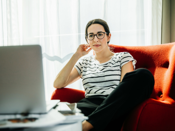 A woman sits in a chair and watches a video on her laptop. 