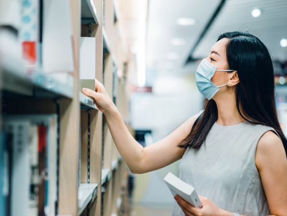 A woman wearing a mask browses books at a library. 